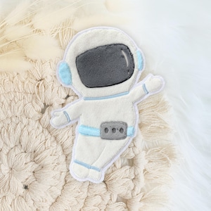 Könnte beinhalten: Weißer Filz-Astronauten-Applikationen mit blauen Details. Der Astronaut trägt einen weißen Raumanzug mit einem blauen Gürtel und einem grauen Helm. Die Applikation befindet sich auf einem weißen Hintergrund.