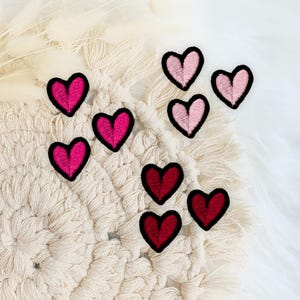 Könnte beinhalten: Eine Ansammlung von herzförmigen, gestickten Aufnähern in verschiedenen Rosa- und Rottönen, jeweils mit schwarzem Rand. Die Aufnäher sind auf einem cremefarbenen Makramee-Untersetzer und einer weißen, flauschigen Oberfläche angeordnet.