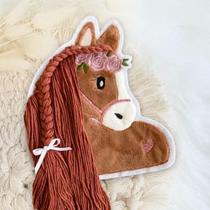 Könnte beinhalten: Ein brauner Plüschpferdekopf mit weißem Gesicht und einem rosa Herzdetail. Das Pferd hat eine rostfarbene Garnmähne, einen Blumenkranz und ein weißes Band. Der Plüsch befindet sich auf einer strukturierten weißen und cremefarbenen Oberfläche.
