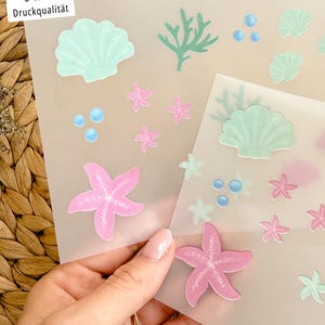 Könnte beinhalten: Blätter mit Aufklebern mit Meeresmotiven. Die Aufkleber umfassen rosa Seesterne, hellblaue Blasen und hellgrüne Muscheln und Korallen. Der Text "B-Ware Druckqualität" und "B-Ware Versatz" ist sichtbar.