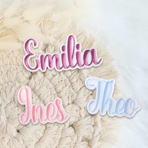 Könnte beinhalten: Drei bestickte Namensaufnäher mit kursiver Schrift. Der obere Aufnäher zeigt den Namen Emilia in einem dunkelrosa Farbverlauf. Der untere linke Aufnäher zeigt den Namen Ines in Hellrosa und der untere rechte Aufnäher den Namen Theo in Hellblau.