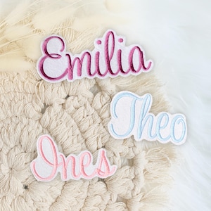 Könnte beinhalten: Drei gestickte Stoff-Patches mit den Namen: Emilia in Rosa, Theo in Hellblau und Ines in Rosa.
