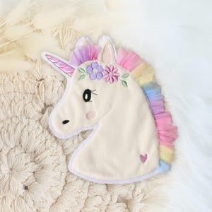 Könnte beinhalten: Cremefarbene Einhorn-Applikation in Kopfform mit Regenbogenmähne und Horn. Das Einhorn hat einen Blumenkranz und ein kleines gesticktes Herz. Die Applikation befindet sich auf einer strukturierten, cremefarbenen Oberfläche.