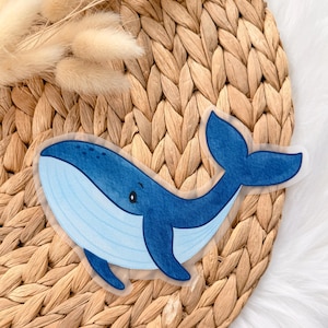 Könnte beinhalten: Ein Aufkleber mit einem blauen Wal mit hellblauem Bauch und dunkelblauen Akzenten. Der Wal hat ein lächelndes Gesicht und befindet sich vor einem gewebten Hintergrund. Der Aufkleber ist ein niedliches, skurriles Design.