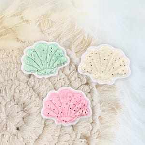 Könnte beinhalten: Drei Filzmuscheln mit goldenen Glitzerakzenten. Die Muscheln sind in den Farben Grün, Rosa und Creme.