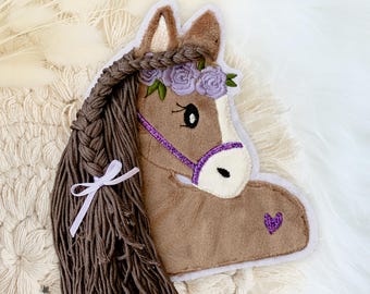 May include: A brown horse head-shaped applique with a purple floral crown and a purple heart. The horse has a long, braided mane with a white ribbon. The applique is on a white, textured surface.
