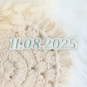 Könnte beinhalten: Ein weißer und grüner gestickter Patch mit dem Datum "11.08.2025" auf einem cremefarbenen Häkelhintergrund.