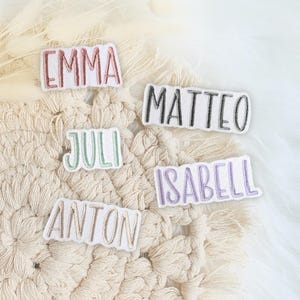 Könnte beinhalten: Fünf weiße Stoffpatches mit gestickten Namen in verschiedenen Farben. Die Namen sind Emma, Matteo, Juli, Anton und Isabell.