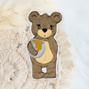 Könnte beinhalten: Ein brauner Plüschbär, der einen Honigtopf hält. Der Bär hat einen weißen Umriss und hält den Topf mit seiner linken Pfote.