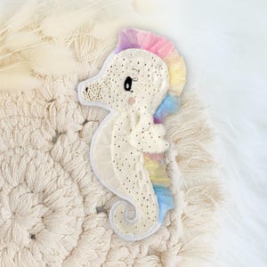 Könnte beinhalten: Ein cremefarbenes Plüschseepferdchen mit einer Regenbogenmähne und -schwanz. Das Seepferdchen hat ein Cartoon-Design mit gestickten Details, darunter kleine schwarze Punkte und ein lächelndes Gesicht. Es liegt auf einem cremefarbenen Makramee-Hintergrund.