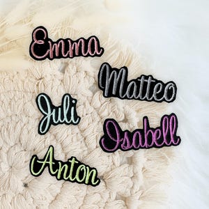 Könnte beinhalten: Gestickte Namensaufnäher in verschiedenen Farben, darunter Rosa, Silber, Grün und Lila, mit schwarzen Rändern. Die Namen sind "Emma", "Matteo", "Juli", "Isabell" und "Anton". Diese Aufnäher sind wahrscheinlich für Kleidung oder Accessoires.
