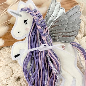 May include: A white felt unicorn with silver wings and a purple and pink braided mane. The unicorn is standing on a white surface.