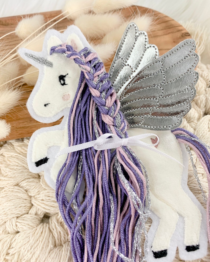 May include: A white felt unicorn with silver wings and a purple and pink braided mane. The unicorn is standing on a white surface.