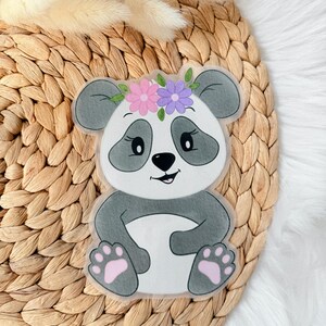 Könnte beinhalten: Ein niedlicher Panda-Aufkleber mit einem Blumenkranz. Der Panda hat eine schwarze Nase, Augen und Ohren mit grauen Akzenten. Seine Pfoten sind rosa. Der Aufkleber befindet sich auf einer gewebten Oberfläche.