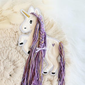 Könnte beinhalten: Ein weißer Einhorn-förmiger Aufnäher mit einem goldenen Horn, schwarzen Hufen und einer geflochtenen lila und goldenen Mähne und Schweif. Ein kleines lila Herz befindet sich an der Seite des Einhorns. Der Aufnäher befindet sich auf einer cremefarbenen, strukturierten Oberfläche.