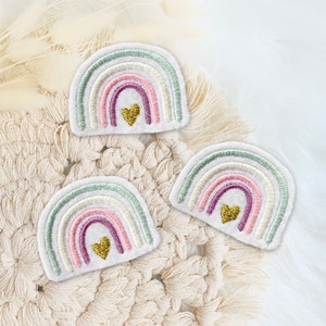Könnte beinhalten: Drei gestickte Regenbogen-Patches mit einem goldenen Herzen in der Mitte jedes Regenbogens. Die Patches befinden sich auf einer weißen gehäkelten Oberfläche.