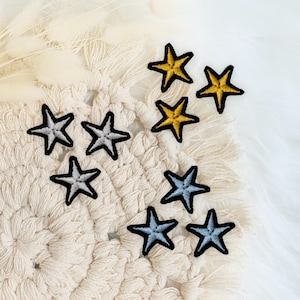 Könnte beinhalten: Gestickte Sternaufnäher in Gold, Silber und Blau mit schwarzen Rändern. Die Aufnäher sind auf einem cremefarbenen Makramee-Untersetzer und einer weißen, flauschigen Oberfläche angeordnet. Ideal zum Aufpeppen von Kleidung oder Accessoires.