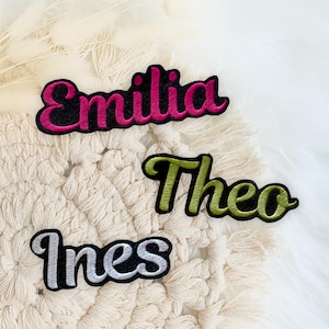 Könnte beinhalten: Drei bestickte Namensaufnäher mit kursiver Schrift und schwarzen Rändern. Der oberste Aufnäher zeigt den Namen Emilia in leuchtendem Pink, der mittlere Aufnäher zeigt Theo in Olivgrün und der untere Aufnäher zeigt Ines in metallischem Silber.