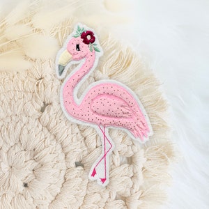 Könnte beinhalten: Rosa-farbener Flamingo-Stickerei-Aufnäher mit einer Blumenkrone. Der Flamingo steht auf einem weißen Makramee-Hintergrund.