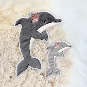 Könnte beinhalten: Zwei graue Filz-Delfin-Applikationen mit rosa Blumen-Stickerei. Der größere Delfin steht aufrecht mit dem Schwanz hinter sich. Der kleinere Delfin steht aufrecht mit dem Schwanz hinter sich. Beide Delfine befinden sich auf einer cremefarbenen gehäkelten Oberfläche.