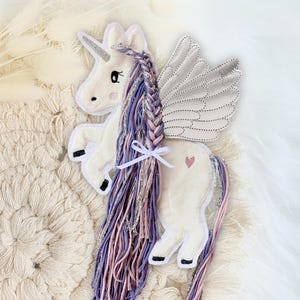 Könnte beinhalten: Ein weißes Plüschtier in Form eines Einhorns mit einem silbernen Glitzerflügel, einer geflochtenen Mähne und einem Schwanz in Lila und Rosa. Das Einhorn hat ein pinkfarbenes Herz an der Seite.