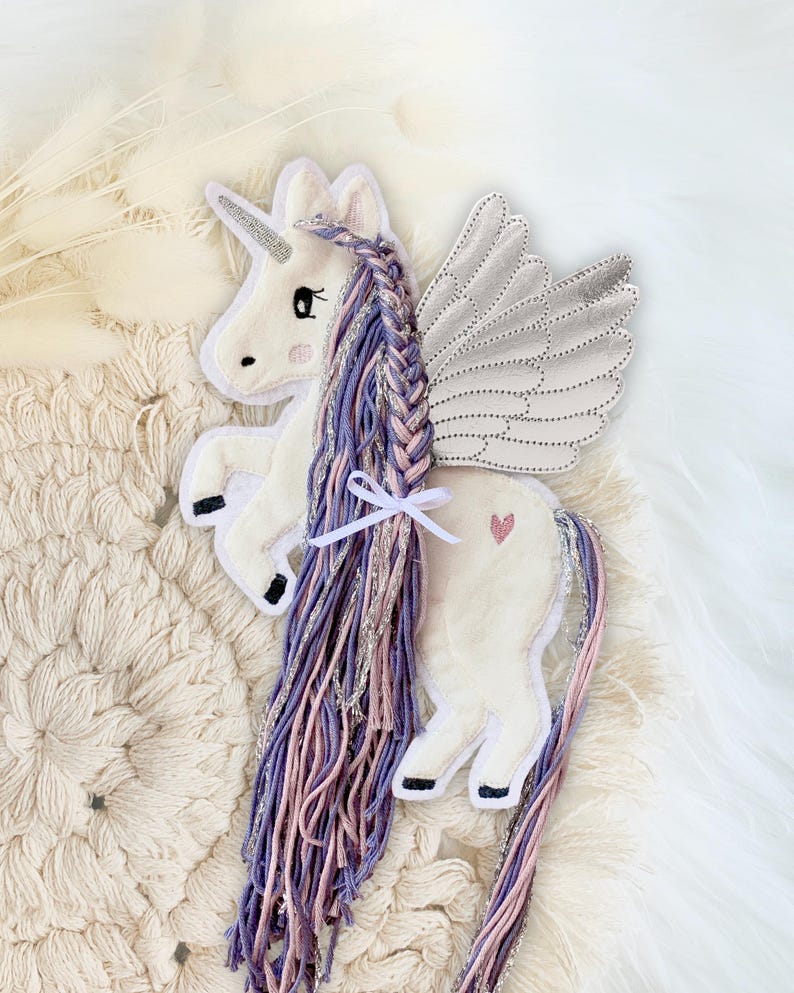 May include: A white unicorn plush toy with a silver glitter wing, purple and pink braided mane and tail. The unicorn has a pink heart on its side.