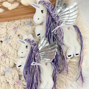 May include: Two felt unicorn ornaments with silver wings and purple and pink yarn hair. The unicorns are standing on a white crocheted surface.