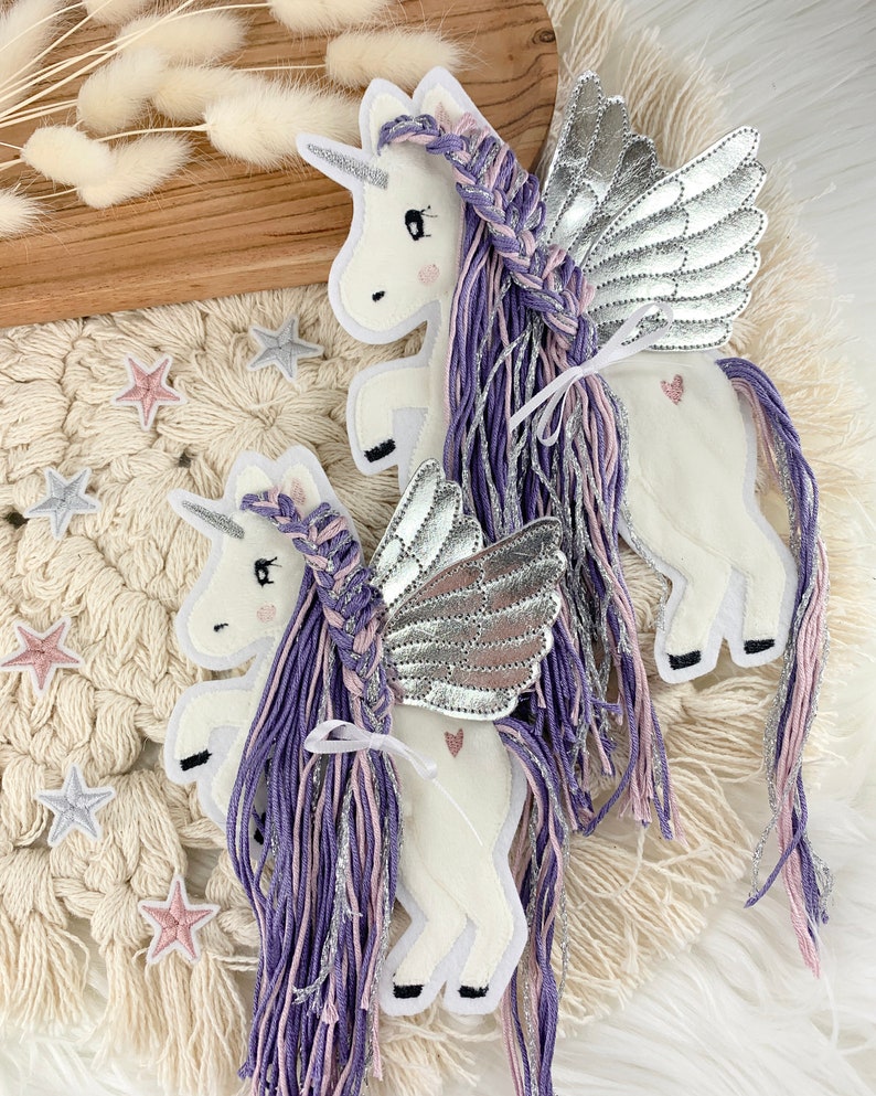 May include: Two felt unicorn ornaments with silver wings and purple and pink yarn hair. The unicorns are standing on a white crocheted surface.