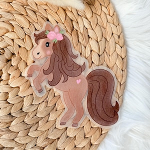 Könnte beinhalten: Ein transparenter Aufkleber mit einem Cartoon-Pony mit braunem Fell, einer dunkelbraunen Mähne und einem Schweif, einem rosa Herz an der Flanke und einem Blumenkranz. Der Aufkleber befindet sich auf einer gewebten, hellbraunen Oberfläche.