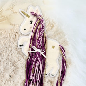 Könnte beinhalten: Weißes Einhorn-Applikation mit goldenem Horn, geflochtener lila und goldener Mähne und einem kleinen goldenen Herz auf der Seite. Das Einhorn hat ein weißes Band um seine Mähne gebunden.