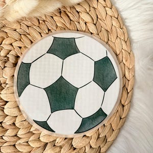 Könnte beinhalten: Ein runder Untersetzer mit einem Fußball-Design. Der Untersetzer hat einen weißen Hintergrund mit schwarzen Fünf- und Sechsecken, die einem Fußball ähneln. Der Untersetzer liegt auf einem gewebten, beigefarbenen Platzset.