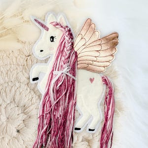 Könnte beinhalten: Ein weißes Filz-Einhorn mit einem rosa Glitzerhorn, einer rosa und weißen geflochtenen Mähne und einem rosa Glitzerschwanz. Das Einhorn hat goldene Flügel und ein kleines rosa Herz an der Seite. Das Einhorn befindet sich auf einer weißen strukturierten Oberfläche.
