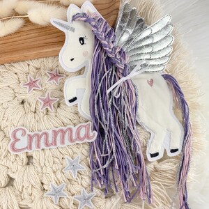 May include: A white unicorn with purple and pink braided hair and silver wings. The unicorn has a pink heart on its side and a pink name tag that says "Emma". There are also several silver and pink stars scattered around the unicorn.