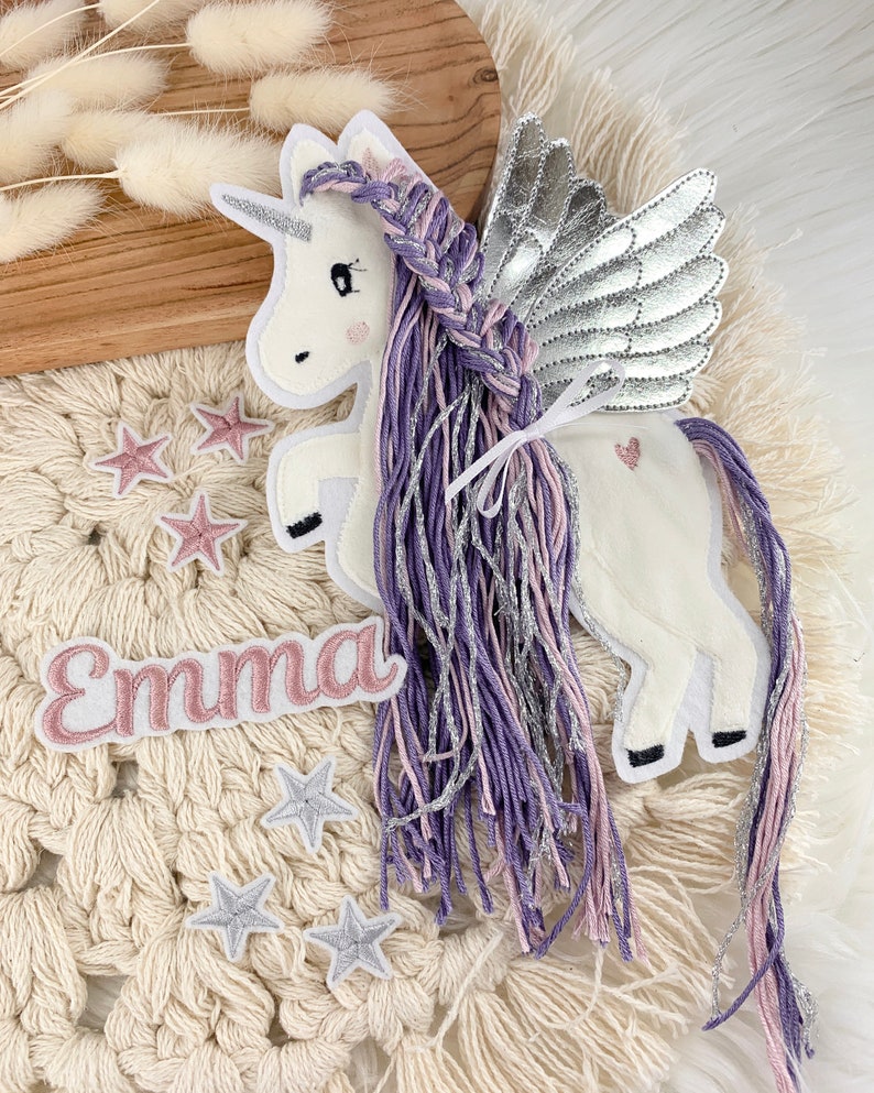 May include: A white unicorn with purple and pink braided hair and silver wings. The unicorn has a pink heart on its side and a pink name tag that says "Emma". There are also several silver and pink stars scattered around the unicorn.