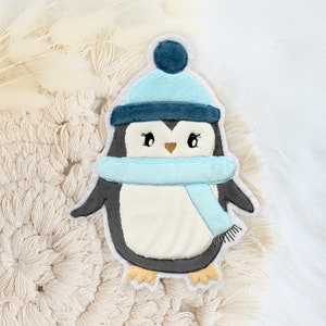Könnte beinhalten: Ein blauer und weißer Pinguin, der einen blauen Hut mit einem weißen Bommel und einen blauen Schal trägt. Der Pinguin befindet sich auf einem weißen Häkelhintergrund.