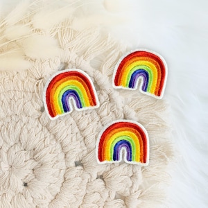 Könnte beinhalten: Drei gestickte Regenbogen-Aufnäher mit roten, orangen, gelben, grünen, blauen und violetten Bögen. Jeder Aufnäher hat einen weißen Rand. Die Aufnäher sind auf einer cremefarbenen, strukturierten Oberfläche mit weißen, flauschigen Akzenten angeordnet.