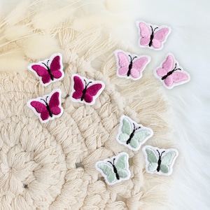 Könnte beinhalten: Eine Sammlung von aufgestickten Schmetterlings-Patches in verschiedenen Farben, darunter Rosa, Magenta und Mintgrün. Jeder Patch zeigt ein detailliertes Schmetterlingsdesign mit schwarzen Fühlern und ist weiß umrandet. Die Patches liegen auf einer strukturierten, cremefarbenen Oberfläche.