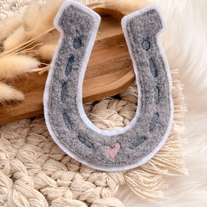 Könnte beinhalten: Ein graues Filz-Hufeisen mit weißem Rand und einem rosa Herz. Das Hufeisen hat dunkelgraue Nahtdetails und kleine, dunkelgraue federartige Formen. Das Hufeisen ist ein Glückssymbol.