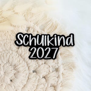 Könnte beinhalten: Ein schwarzer Stickaufnäher mit weißer gestickter Schrift, die Schulkind 2027 lautet. Der Aufnäher hat eine kräftige, abgerundete Schriftart und liegt auf einem kreisförmigen, cremefarbenen Makramee-Textil mit Fransenrand und getrocknetem Gras.