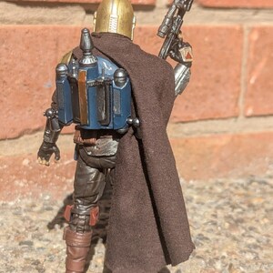 K&ouml;nnte beinhalten: Detaillierte Actionfigur einer Figur aus einem Science-Fiction-Franchise, von hinten gesehen. Die Figur tr&auml;gt einen goldenen Helm, einen braunen Umhang und eine blaue und silberne R&uuml;stung. Sie h&auml;lt ein schwarzes Gewehr mit Zielfernrohr. Die Figur steht auf einer Steinoberfl&auml;che.