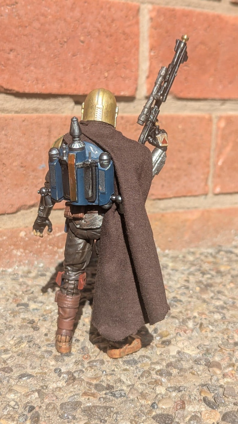 K&ouml;nnte beinhalten: Detaillierte Actionfigur einer Figur aus einem Science-Fiction-Franchise, von hinten gesehen. Die Figur tr&auml;gt einen goldenen Helm, einen braunen Umhang und eine blaue und silberne R&uuml;stung. Sie h&auml;lt ein schwarzes Gewehr mit Zielfernrohr. Die Figur steht auf einer Steinoberfl&auml;che.