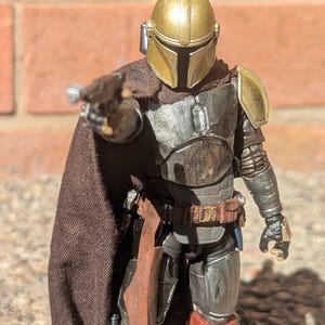 K&ouml;nnte beinhalten: Detaillierte Actionfigur eines Charakters mit goldenem Helm und dunkler R&uuml;stung, der eine Waffe zeigt. Die Figur hat einen braunen Umhang und posiert vor einer Backsteinmauer. Die R&uuml;stung der Figur ist metallfarben.