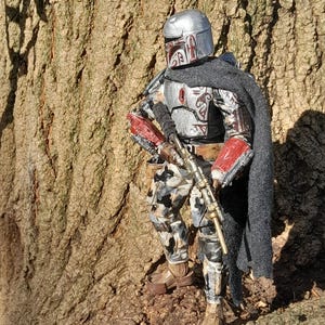 K&ouml;nnte beinhalten: Eine silberne und rote Actionfigur eines Kopfgeldj&auml;gers mit Helm und Gewehr. Die Figur steht vor einem Baumstamm.