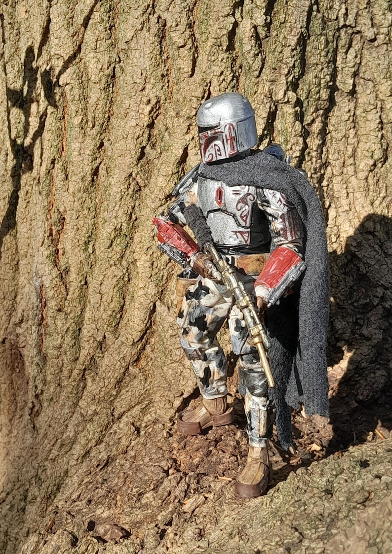 K&ouml;nnte beinhalten: Eine silberne und rote Actionfigur eines Kopfgeldj&auml;gers mit Helm und Gewehr. Die Figur steht vor einem Baumstamm.