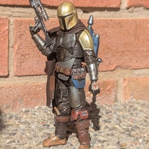 K&ouml;nnte beinhalten: Detaillierte Actionfigur eines Mandalorianischen Kriegers, gekleidet in dunkler R&uuml;stung mit goldenen Akzenten auf Helm und Schulterpolstern. Die Figur h&auml;lt ein Blastergewehr und hat einen Jetpack. Die Figur steht auf einer Steinoberfl&auml;che.