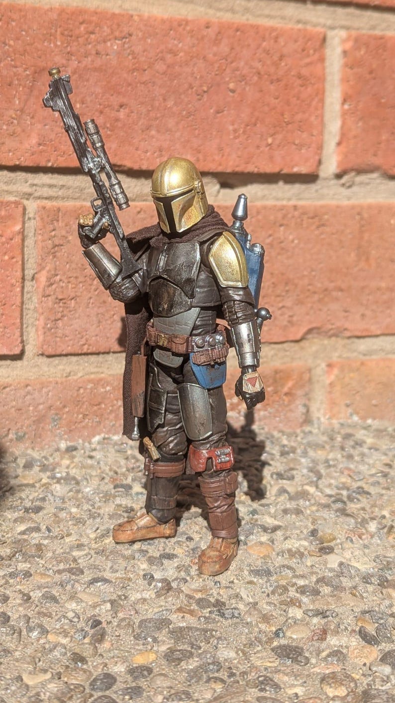 K&ouml;nnte beinhalten: Detaillierte Actionfigur eines Mandalorianischen Kriegers, gekleidet in dunkler R&uuml;stung mit goldenen Akzenten auf Helm und Schulterpolstern. Die Figur h&auml;lt ein Blastergewehr und hat einen Jetpack. Die Figur steht auf einer Steinoberfl&auml;che.