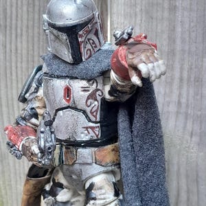 K&ouml;nnte beinhalten: Eine verwitterte, wei&szlig;e und silberne Actionfigur eines Mandalorianer-Kriegers mit einem rot-schwarzen Helm, der eine Blasterpistole h&auml;lt. Die Figur tr&auml;gt einen grauen Umhang und hat ein rot-schwarzes Design auf der Brustpanzerung.