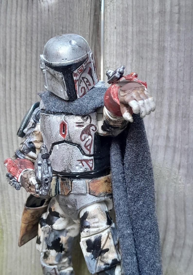 K&ouml;nnte beinhalten: Eine verwitterte, wei&szlig;e und silberne Actionfigur eines Mandalorianer-Kriegers mit einem rot-schwarzen Helm, der eine Blasterpistole h&auml;lt. Die Figur tr&auml;gt einen grauen Umhang und hat ein rot-schwarzes Design auf der Brustpanzerung.
