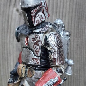 K&ouml;nnte beinhalten: Eine silberne und rote Mandalorianer-Actionfigur mit einem verwitterten und abgenutzten Aussehen. Die Figur tr&auml;gt einen Helm mit einem roten und wei&szlig;en Design und einen Jetpack. Die Figur h&auml;lt ein Blastergewehr in der rechten Hand.