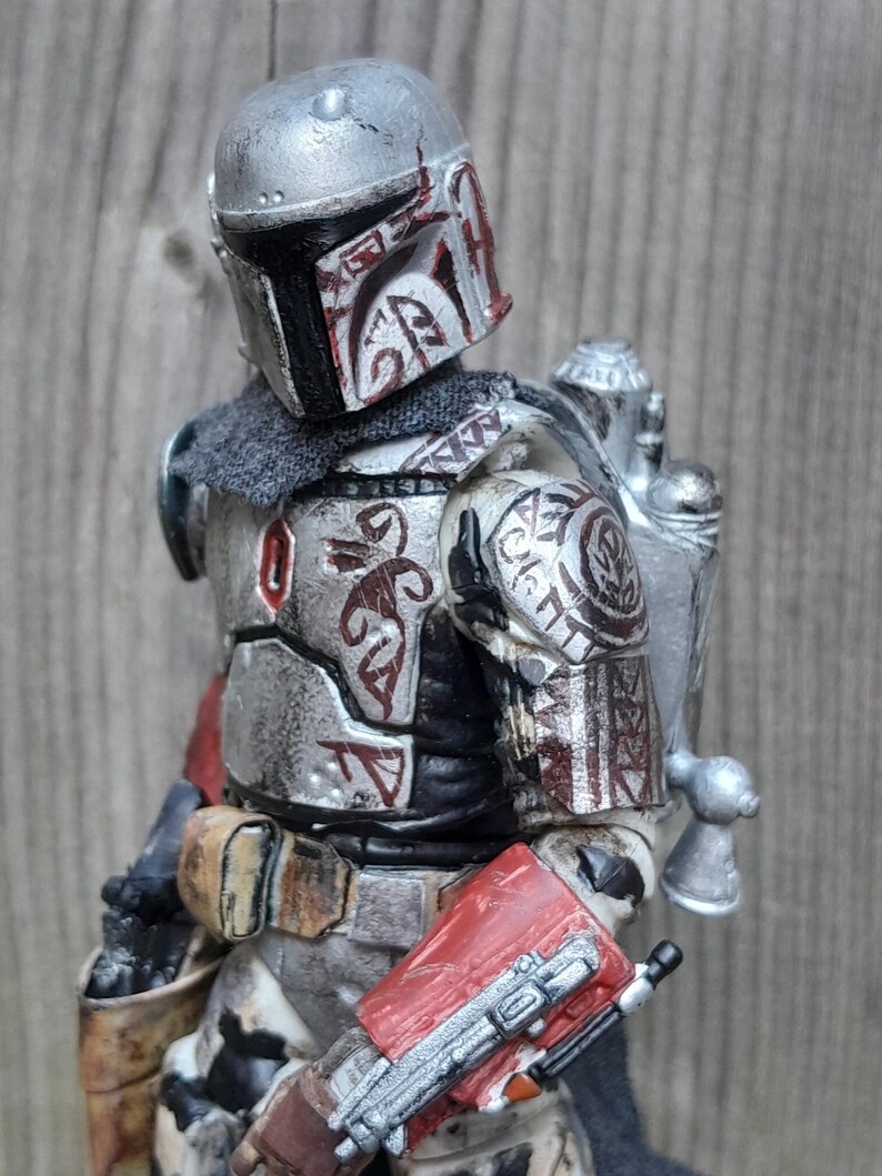 K&ouml;nnte beinhalten: Eine silberne und rote Mandalorianer-Actionfigur mit einem verwitterten und abgenutzten Aussehen. Die Figur tr&auml;gt einen Helm mit einem roten und wei&szlig;en Design und einen Jetpack. Die Figur h&auml;lt ein Blastergewehr in der rechten Hand.